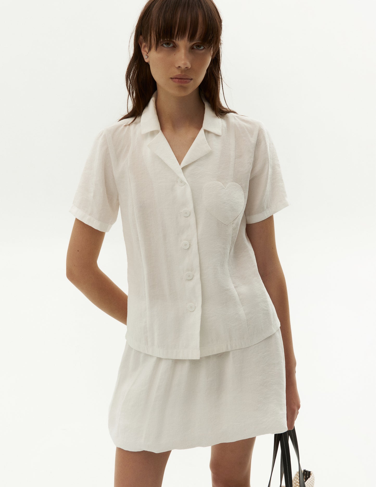 stunning white shirt, featuring a chic lapel collar, short sleeves, and a straight hem. The shirt is designed with a unique twist, featuring a chest heart patch pocket for added style. With a flattering side slit and a front button fastening, this shirt is perfect for any occasion. Match this with our viscose white balloon skirt and wear it like a suit for a stylish and coordinated look, FORMA fashion brand